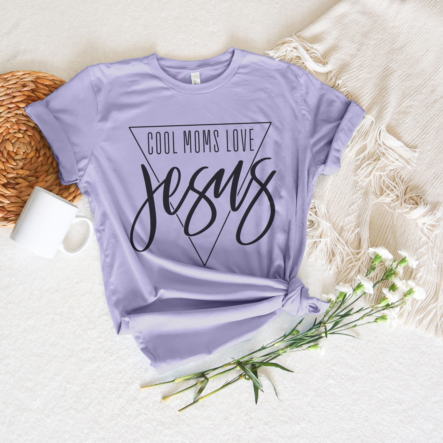 The Cool Moms Love Jesus short sleeve tee, a lavender shirt made from buttery soft fabric with its slogan boldly in a triangle, rests on a cream fringed blanket. It’s complemented by a mug, rattan placemat, and white flowers, offering a perfect blend of comfort and style for believers.
