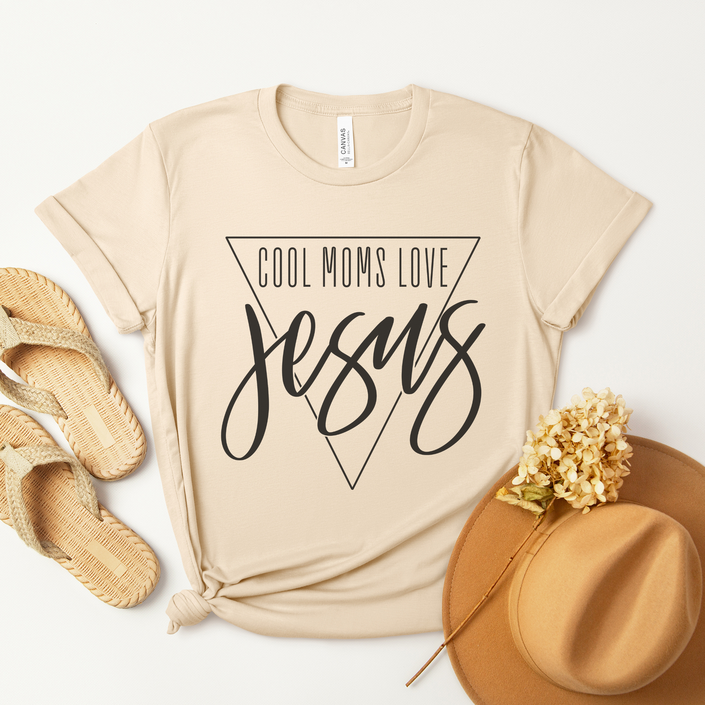 The Cool Moms Love Jesus Short Sleeve Tee is made from buttery soft beige Airlume combed cotton, featuring the phrase in bold script inside an inverted triangle. Styled with a bottom knot, it pairs well with woven sandals and a brown hat amid dried flowers on a white background.