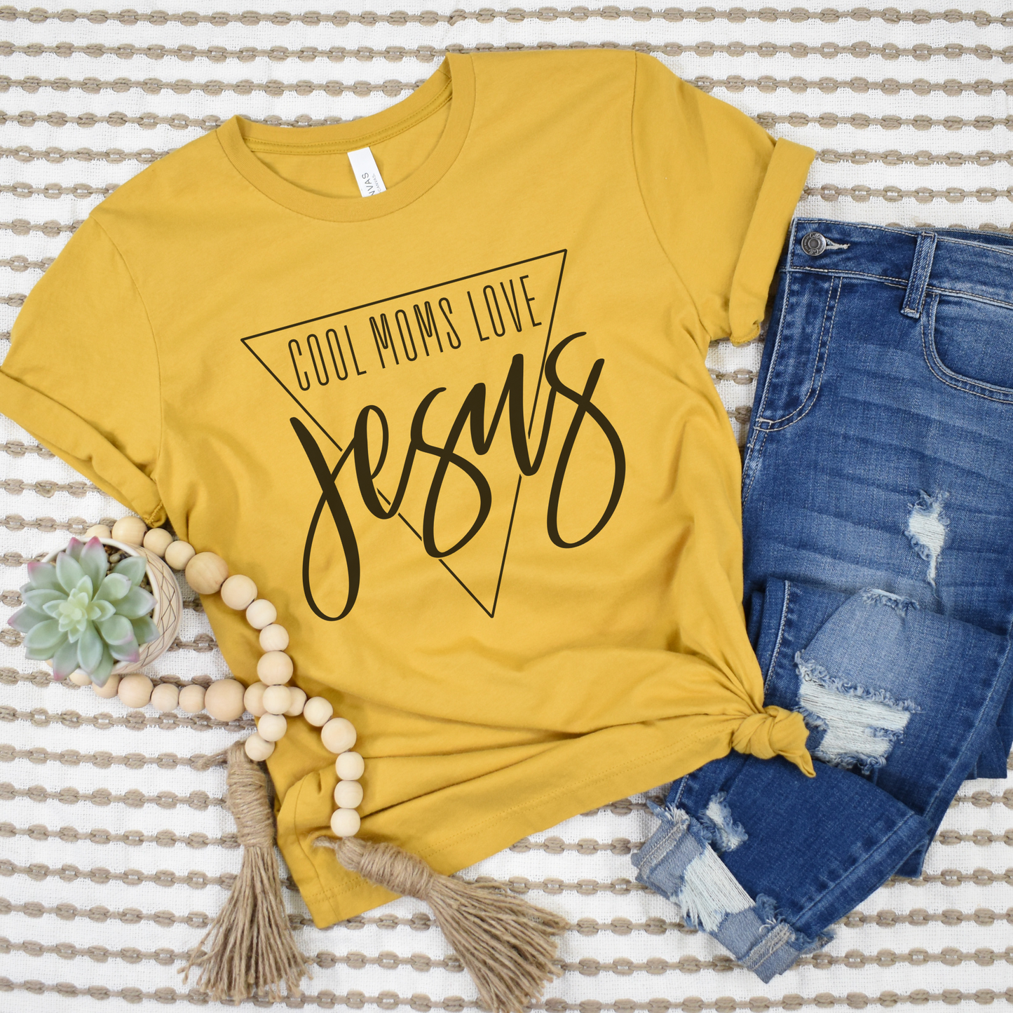 A buttery soft yellow Cool Moms Love Jesus Short Sleeve Tee lies on a striped background. Made from Airlume combed cotton, its paired with ripped blue jeans, a wooden bead bracelet with tassels, and a small succulent plant to complete the look.
