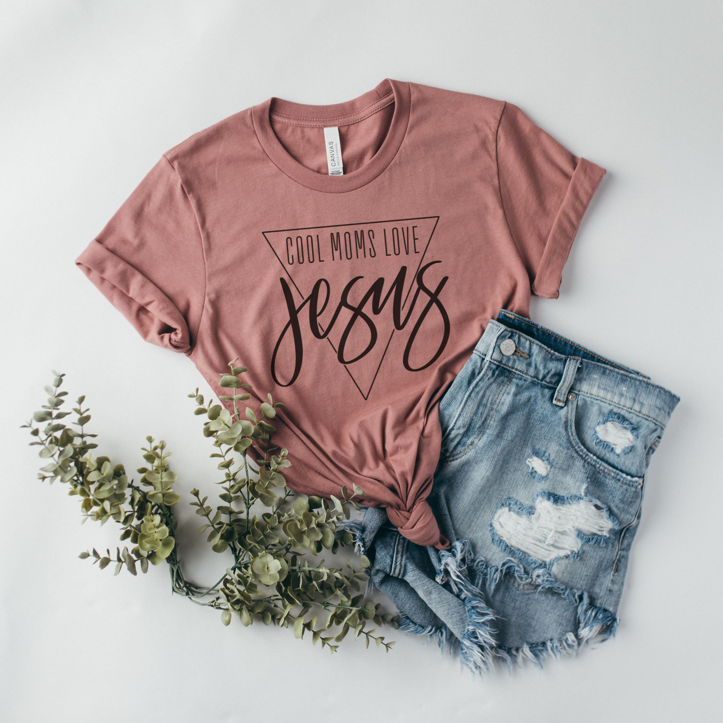 The Cool Moms Love Jesus Short Sleeve Tee, in pink and crafted from buttery soft Airlume combed cotton, is laid flat with ripped denim shorts on a white surface, accented with green foliage for decoration.