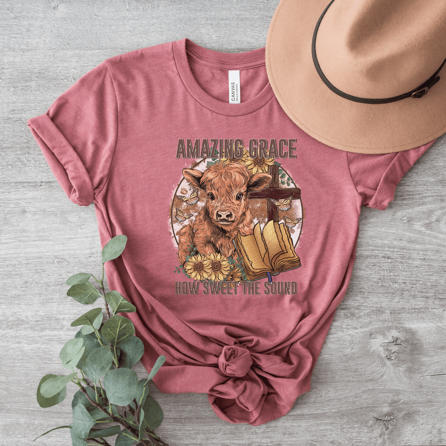 A circle with a highland calf, sunflowers, a cross, and an open bible with the words "Amazing Grace" above and "How Sweet The Sound" below. Design is on a mauve shirt.