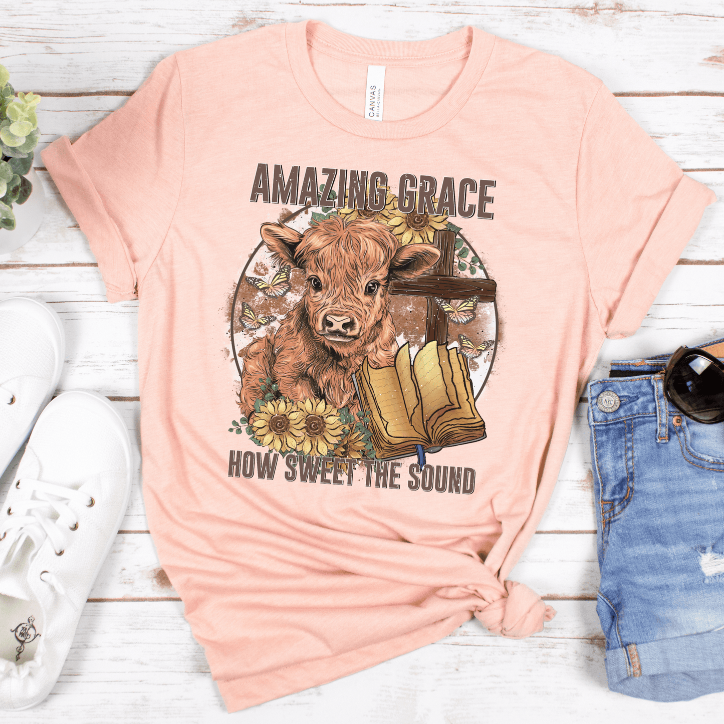 A circle with a highland calf, sunflowers, a cross, and an open bible with the words "Amazing Grace" above and "How Sweet The Sound" below. Design is on a peach colored shirt.