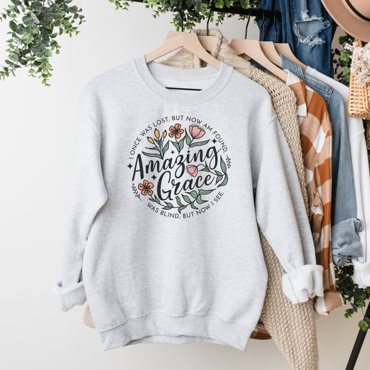 Words "Amazing Grace" with florals around and "I once was lost but now am found, was blind but now I see" in a circle around the main design. Design is on an ash colored sweatshit hanging on a rack.
