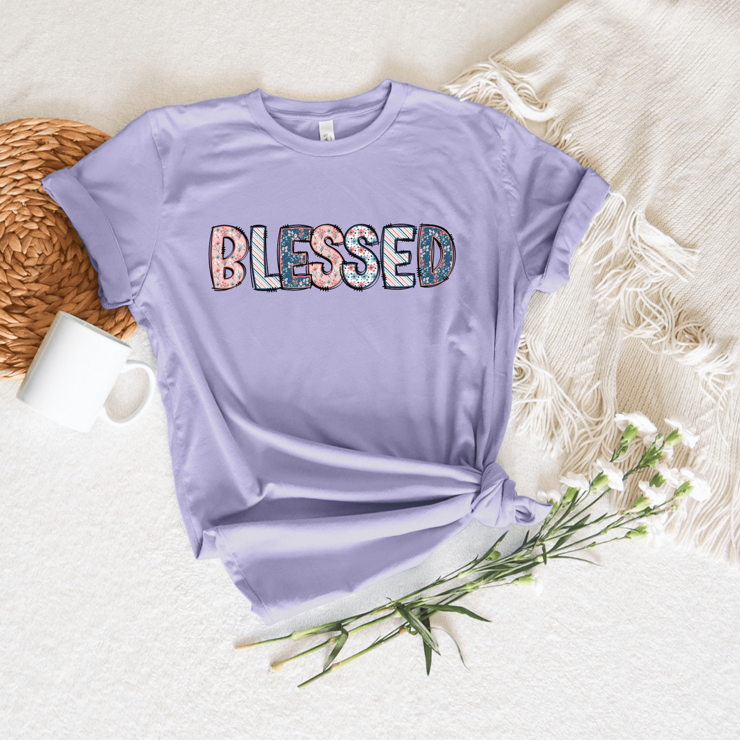 The Blessed Short Sleeve Tee in lilac, made from buttery soft fabric, features BLESSED in floral-patterned letters and is styled knotted at the front. Its placed on a cream blanket alongside a white mug, woven placemat, and white flowers for an inspiring display.