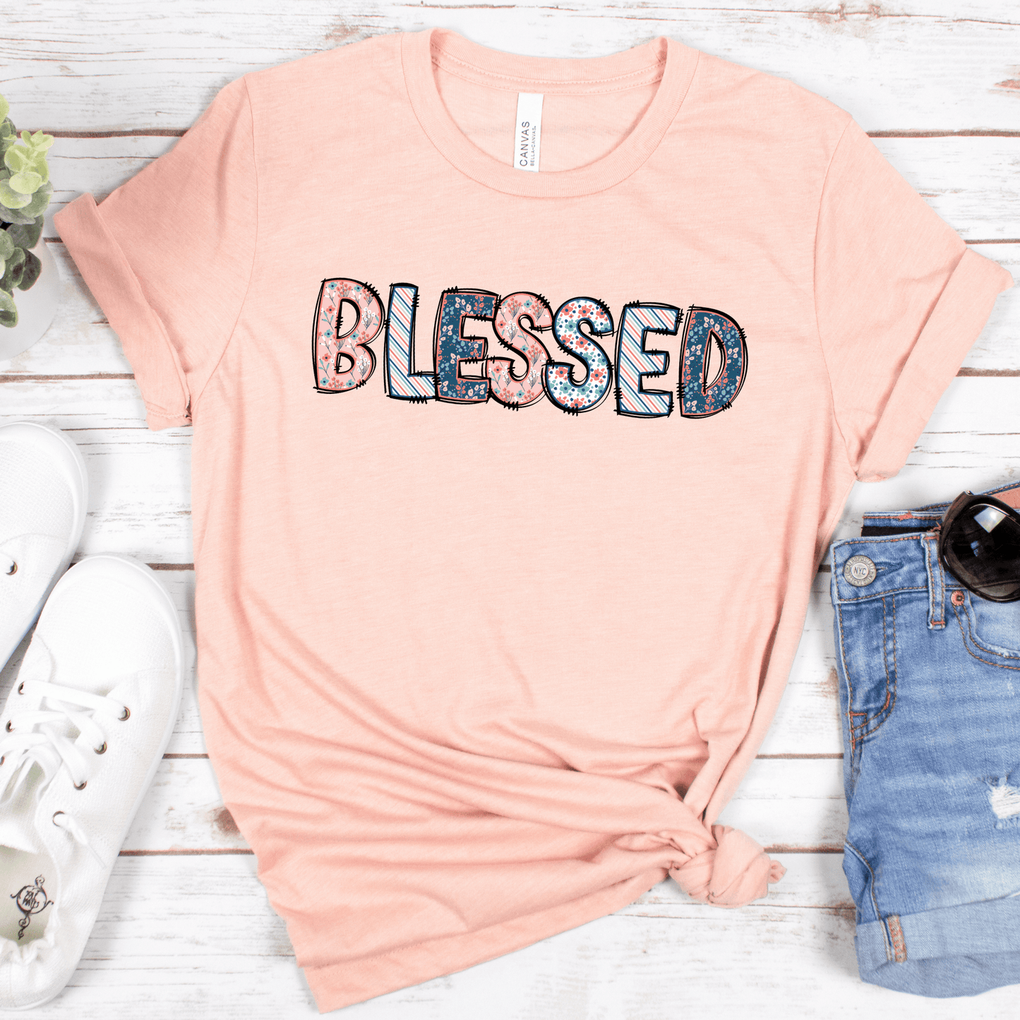 The Blessed Short Sleeve Tee, crafted from buttery soft Airlume combed cotton, features BLESSED in colorful letters. Styled on a white wooden surface, it pairs well with white sneakers, distressed denim shorts, sunglasses, and a small potted plant.