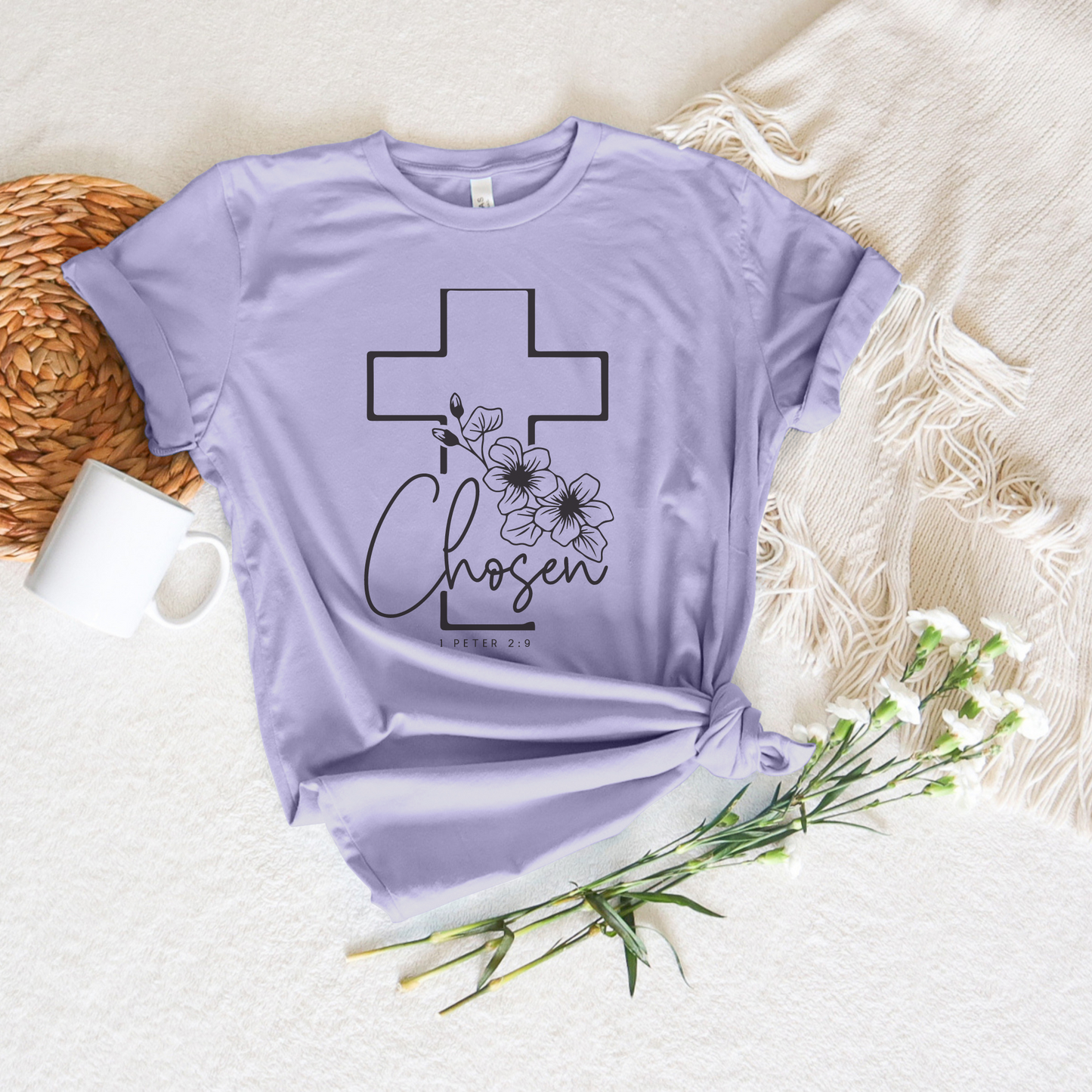 The Chosen Short Sleeve Tee in light purple is made from soft Airlume cotton and features a cross, floral design, and Chosen, 1 Peter 2:9 text. Its stylishly paired with a beige fringe scarf, white mug, wicker placemat, and white flowers. Designed with God in mind.