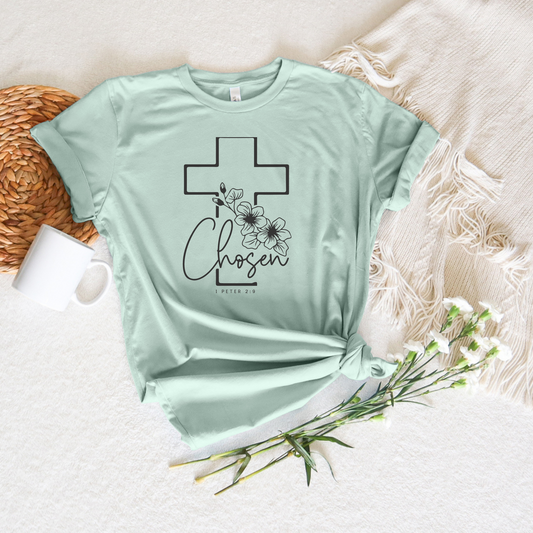 The Chosen Short Sleeve Tee in light green, made from soft Airlume cotton, features a cross and floral design with Chosen and I Peter 2:9. Displayed on a cream blanket near a woven mat, white mug, and small white flowers, its designed with God in mind.