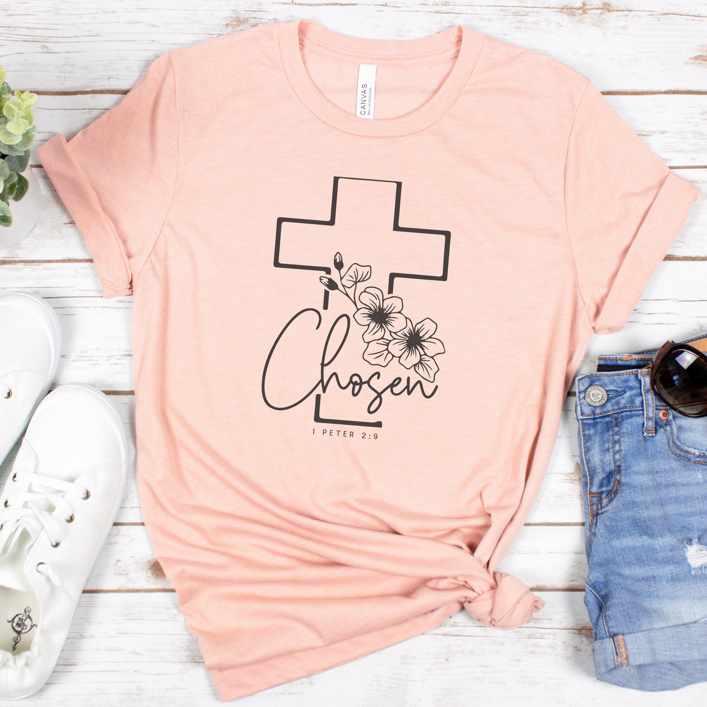 The Chosen Short Sleeve Tee is a buttery soft pink T-shirt made from Airlume cotton, featuring a black cross design with floral accents and the text Chosen and 1 Peter 2:9, styled flat with white sneakers, sunglasses, denim shorts, and an artfully arranged small plant.