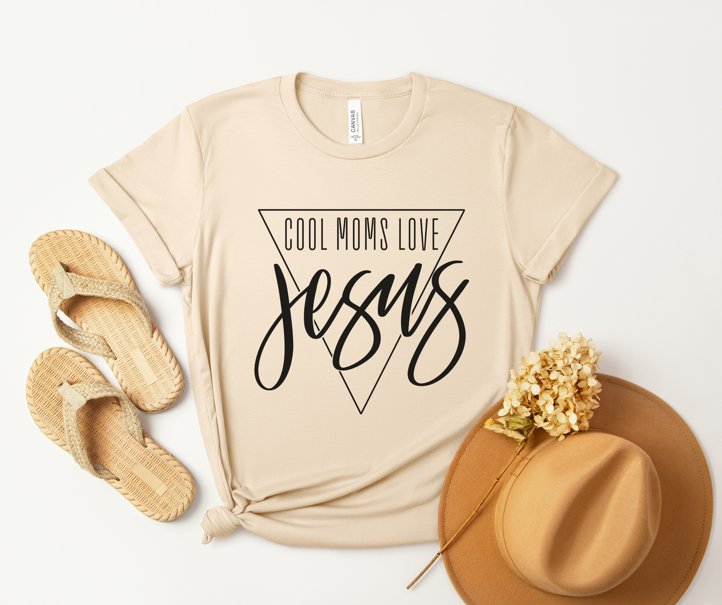 The Cool Moms Love Jesus Short Sleeve Tee, crafted from soft Airlume combed cotton in beige, showcases the phrase inside a triangle with a knotted hem. Its styled alongside woven sandals, a wide-brimmed hat, and an artistic arrangement of dried flowers.