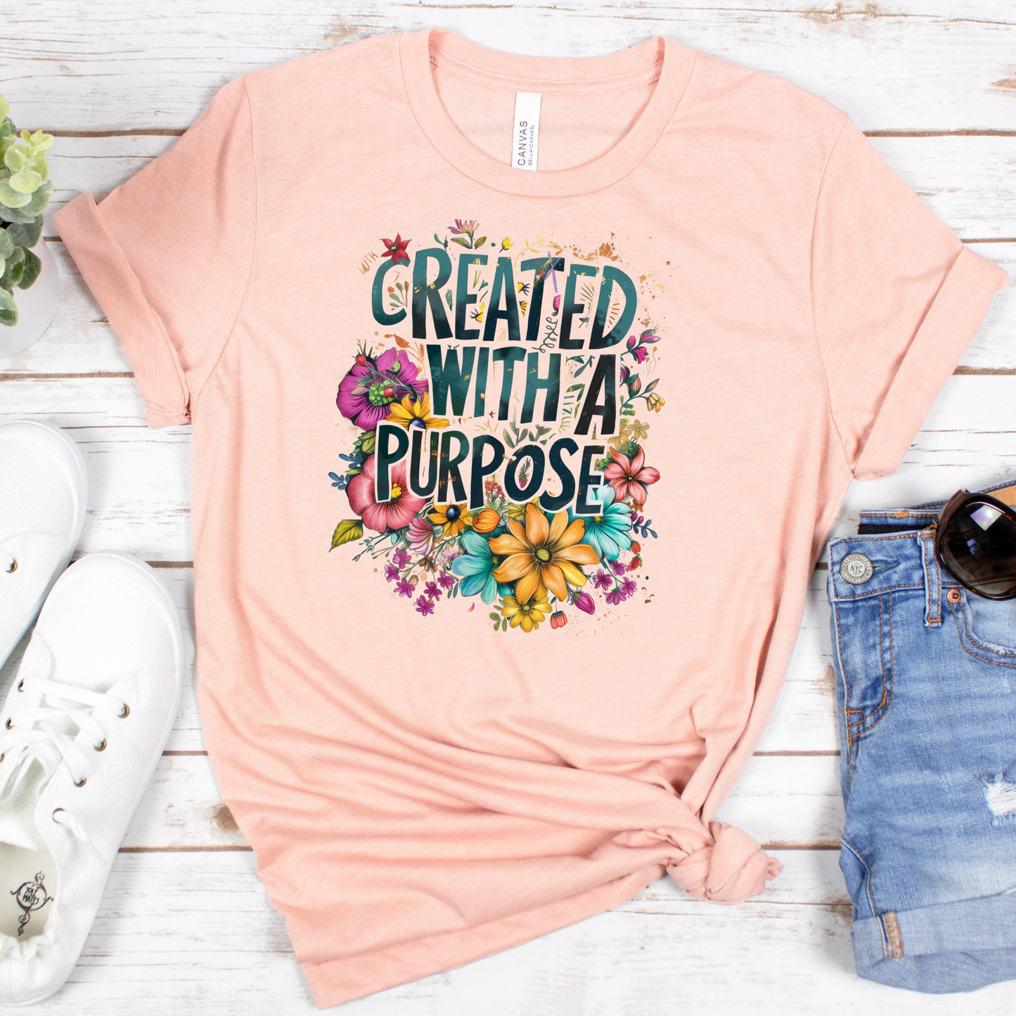 The Created With A Purpose Floral Style Short Sleeve Tee is peach-colored with multicolored flowers on buttery soft Airlume combed cotton, resting against a white rustic wood backdrop with sneakers, denim, and sunglasses.