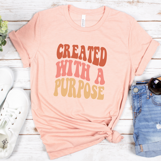 The Created With A Purpose Retro Style Short Sleeve Tee is a peach-colored, buttery soft Airlume cotton shirt with the phrase CREATED WITH A PURPOSE in bold, multicolored letters. Pictured on a wooden surface with white sneakers, denim shorts, and sunglasses. Designed perfectly with God in mind.