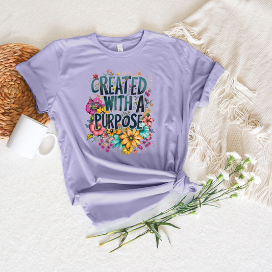 The Created With A Purpose Floral Style short sleeve tee in soft lavender, adorned with colorful flowers and text, rests on a cream blanket beside a white mug, woven placemat, and white flowers, capturing a God-inspired design.