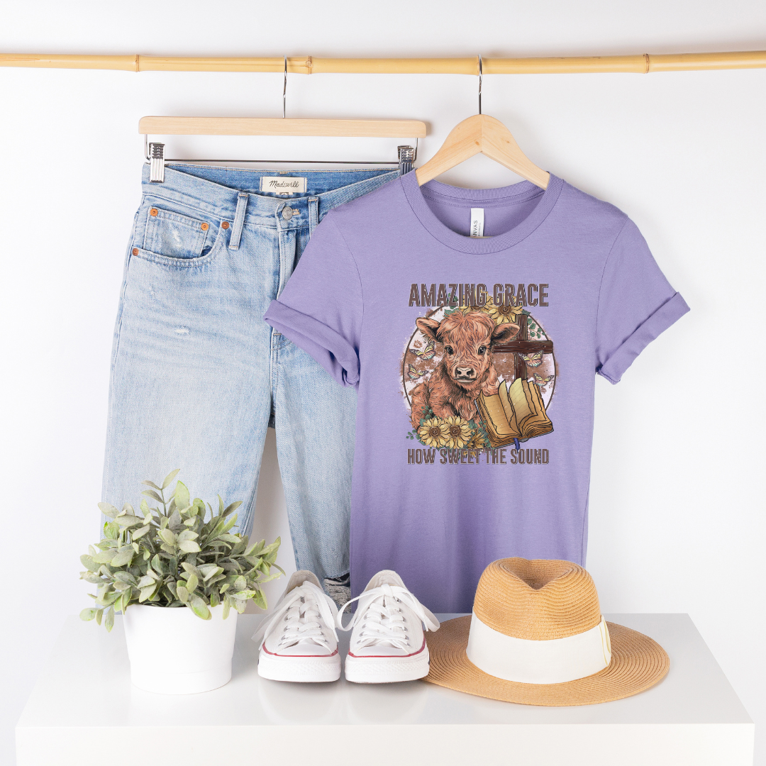 A circle with a highland calf, sunflowers, a cross, and an open bible with the words "Amazing Grace" above and "How Sweet The Sound" below. Design is on a light purple shirt.