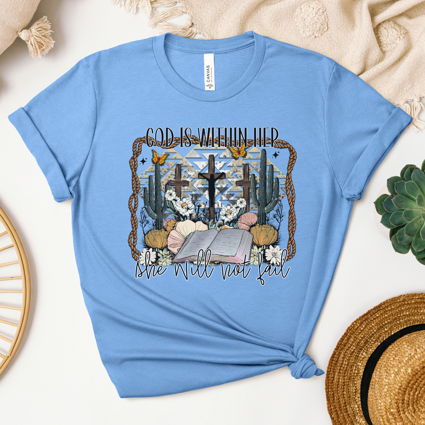 The God Is Within Her Western Short Sleeve Tee is a blue t-shirt made from soft Airlume cotton, featuring the text God Is Within Her, She Will Not Fall alongside designs of mountains, flowers, seashells, cacti, and birds. Displayed on textured surface with greenery and woven hat nearby.