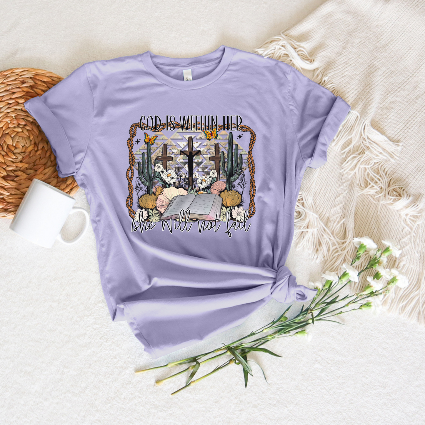 Lilac God Is Within Her Western Short Sleeve Tee made of buttery soft Airlume cotton with a floral cross design and the text, “God is within her, she will not fail. Styled flat with a woven placemat, white fringe blanket, bouquet, and mug. Designed perfectly with God in mind.