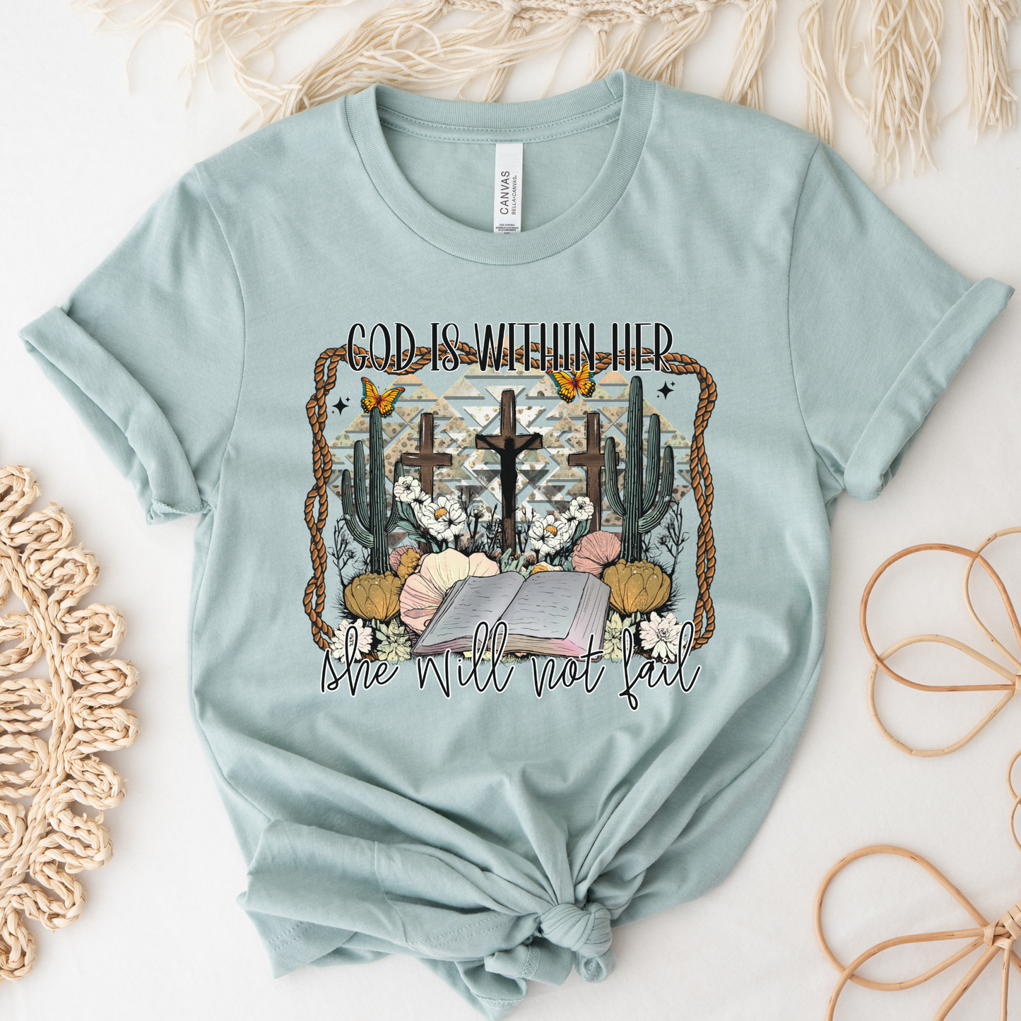 The God Is Within Her Western Short Sleeve Tee is crafted from soft Airlume cotton in light blue, featuring a desert scene with crosses, cacti, and flowers. It reads God is within her above and she will not fail below, enhanced with natural accents for a serene appeal.
