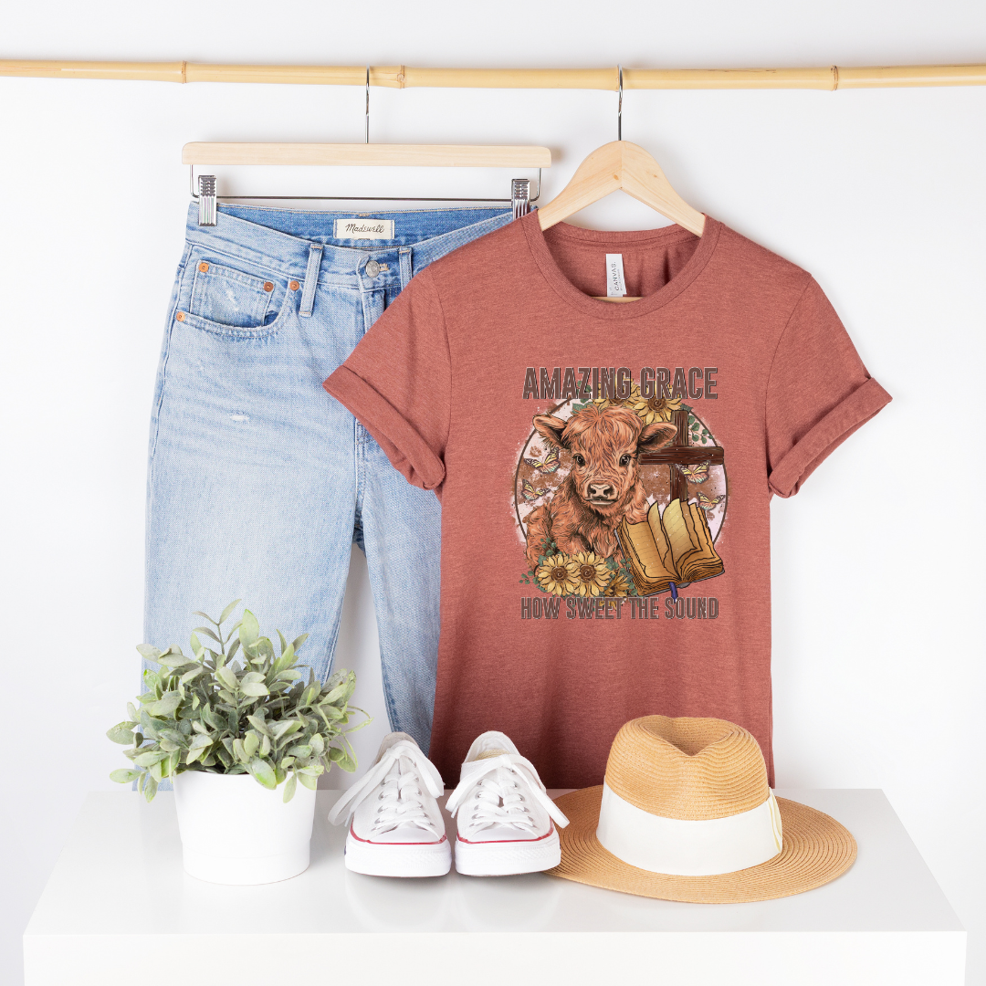 A circle with a highland calf, sunflowers, a cross, and an open bible with the words "Amazing Grace" above and "How Sweet The Sound" below. Design is on a heather orange shirt.