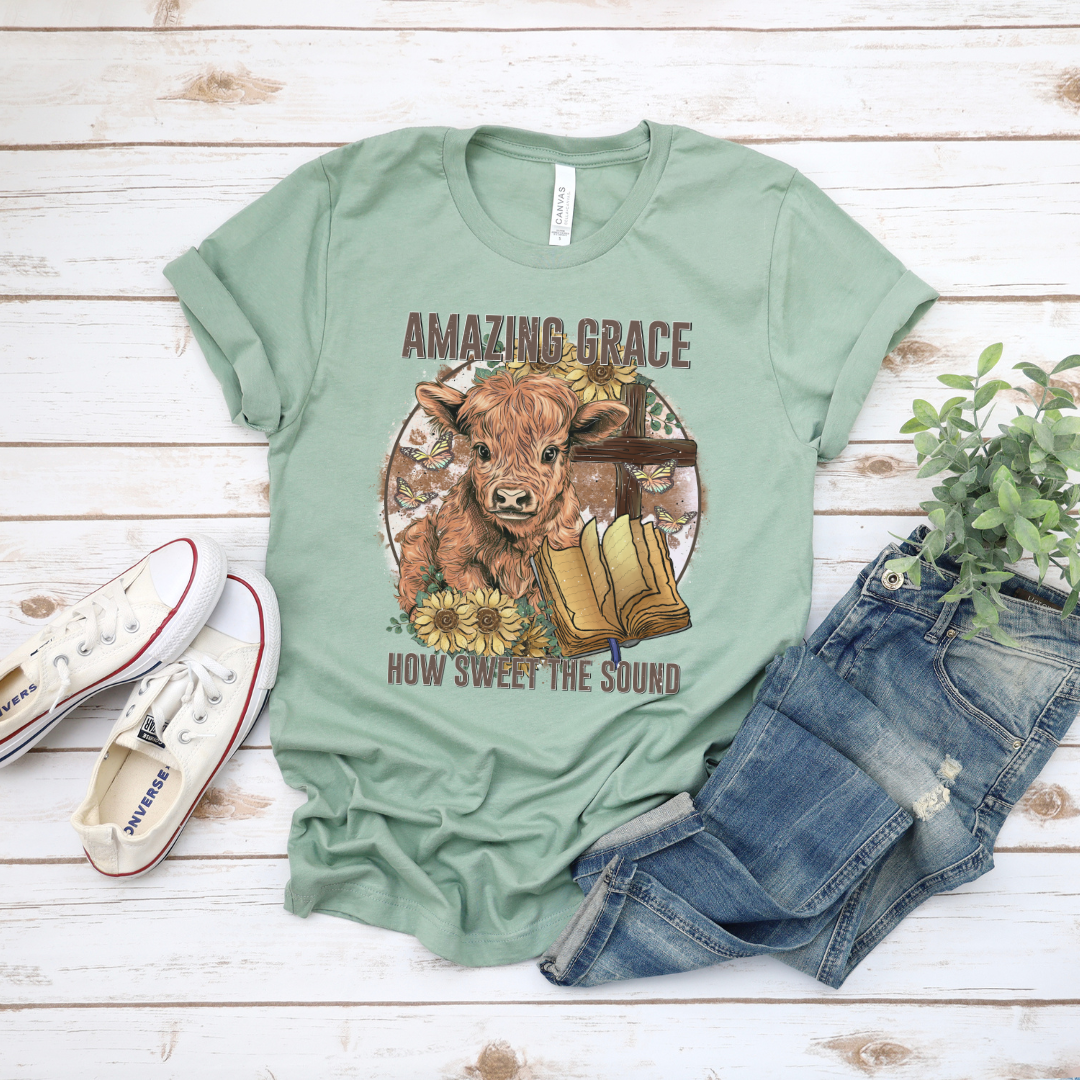 A circle with a highland calf, sunflowers, a cross, and an open bible with the words "Amazing Grace" above and "How Sweet The Sound" below. Design is on a light sage colored shirt.