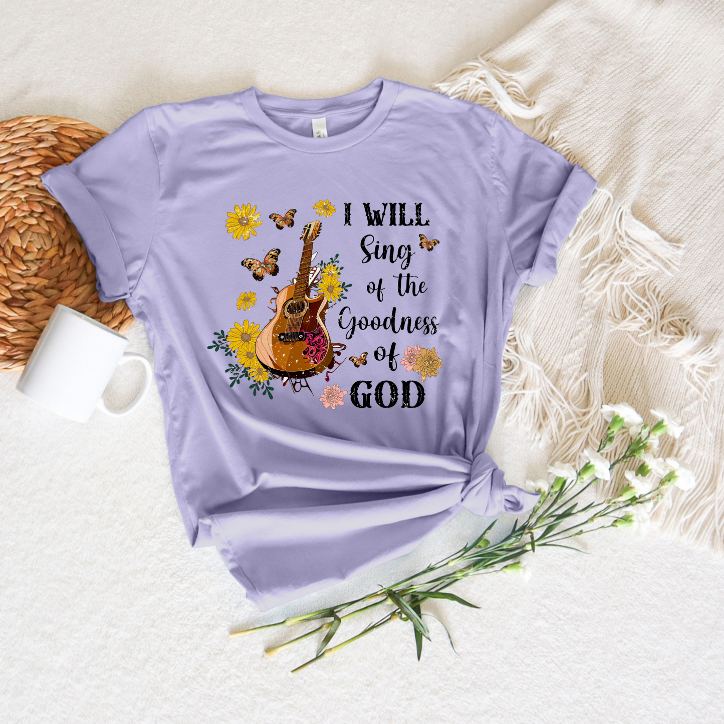 The I Will Sing of The Goodness of God short sleeve tee, in buttery soft lavender Airlume cotton, displays a guitar graphic with flowers and butterflies, accompanied by the text I will sing of the goodness of God, styled next to a woven mat, mug, fringed cloth, and white flowers.