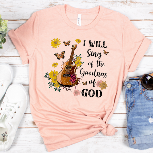 The I Will Sing of The Goodness of God Short Sleeve Tee is buttery soft in pastel pink, featuring a guitar with flowers and butterflies. The text reads, I Will Sing of the Goodness of God. Nearby lie white sneakers and blue denim shorts. Its made from 100% Airlume cotton for ultimate comfort.