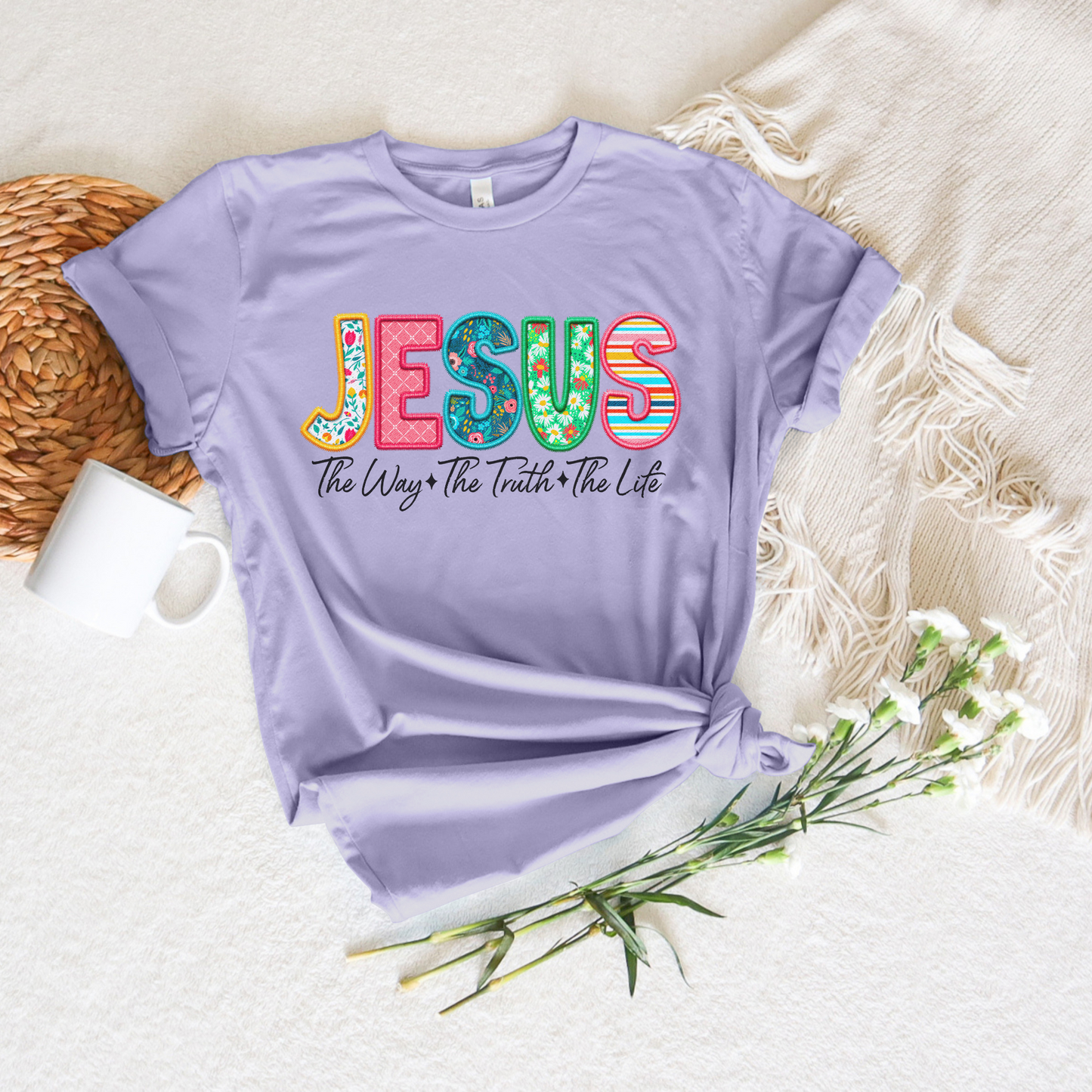 This buttery soft lavender tee, crafted from Airlume cotton, showcases bold letters spelling JESUS with The Way The Truth The Life below. Styled alongside a white fringed mat, a mug, woven coaster, and white flowers. Product: Jesus The Way The Truth The Life (Spring Floral) Short Sleeve Tee.