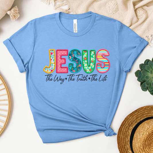 The Jesus The Way The Truth The Life (Spring Floral) short sleeve tee features light blue Airlume cotton with vibrant JESUS lettering and text beneath. Styled knotted at the bottom right, it sits on a textured surface beside a straw hat and plant, exuding true craftsmanship.