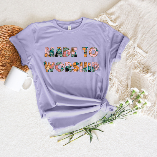 The Made to Worship Tropical Floral Short Sleeve Tee, crafted from buttery soft Airlume combed cotton in lavender, showcases vibrant floral letters. Its beautifully arranged on a beige blanket alongside a white cup, woven placemat, and delicate white flowers.