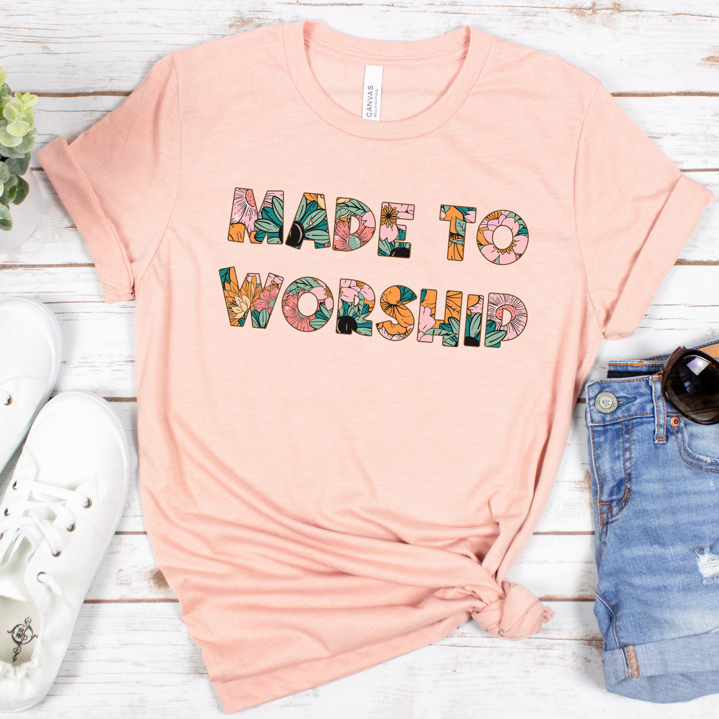 The Made to Worship Tropical Floral Short Sleeve Tee is buttery soft and in Airlume combed cotton. Styled chicly with white sneakers, a small potted plant, denim shorts, and sunglasses on a rustic white wooden surface, it features colorful floral letters.