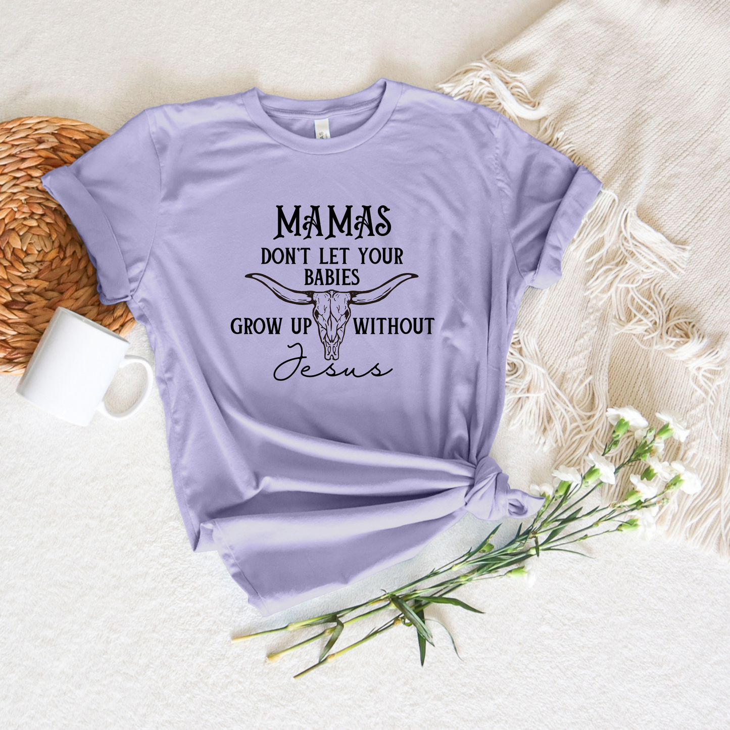 Lilac Mamas Dont Let Your Babies Grow Up Without Jesus short sleeve tee with a bull skull design on a textured backdrop, paired with a white mug, woven basket, and delicate white flowers.