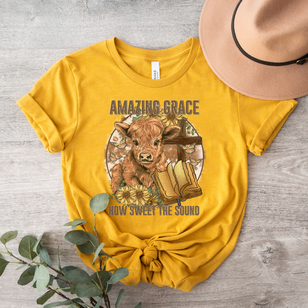 A circle with a highland calf, sunflowers, a cross, and an open bible with the words "Amazing Grace" above and "How Sweet The Sound" below. Design is on a mustard colored shirt.