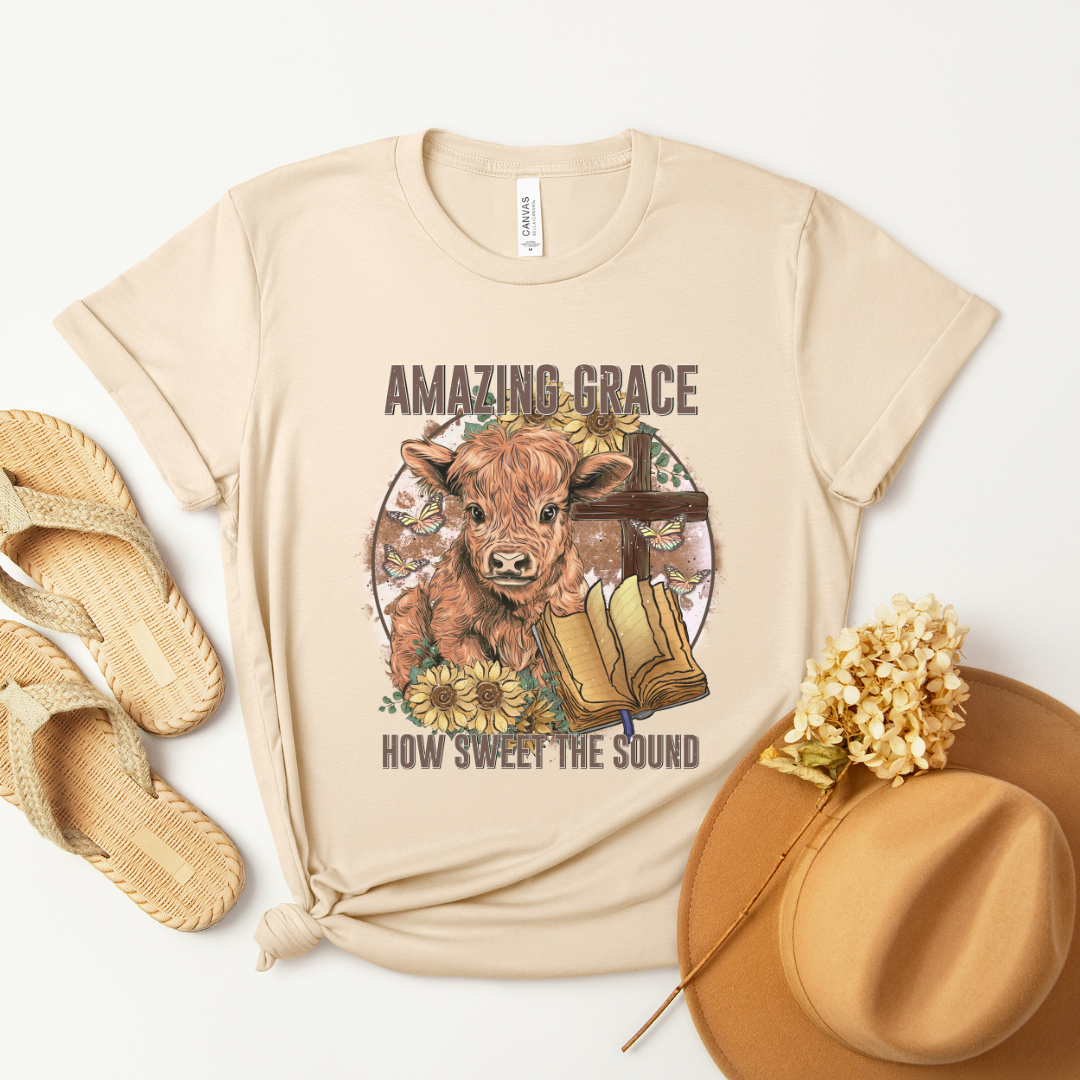 A circle with a highland calf, sunflowers, a cross, and an open bible with the words "Amazing Grace" above and "How Sweet The Sound" below. Design is on a light tan shirt.