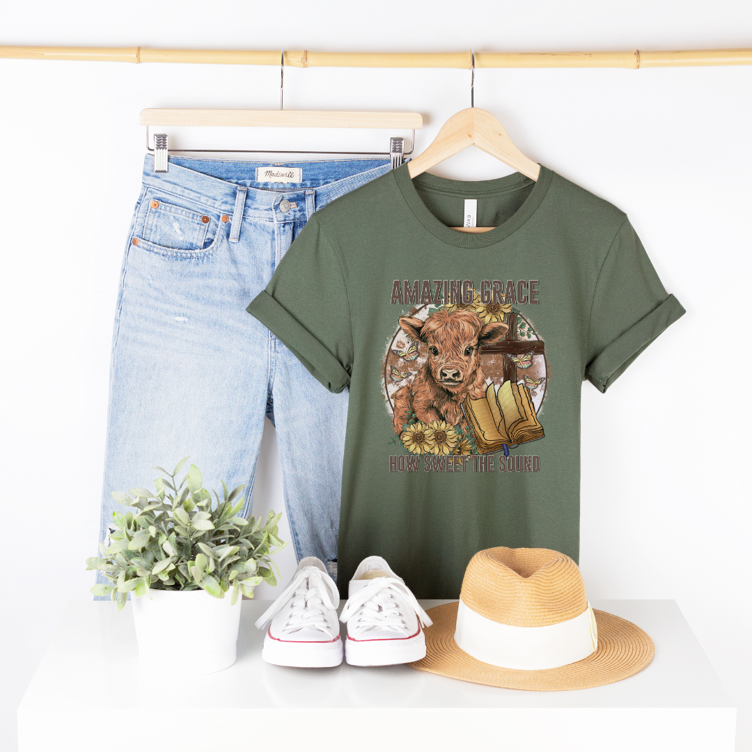 A circle with a highland calf, sunflowers, a cross, and an open bible with the words "Amazing Grace" above and "How Sweet The Sound" below. Design is on a pine colored shirt.