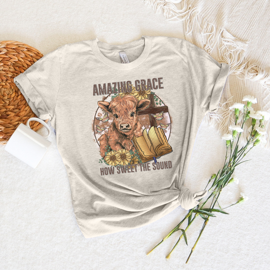 A circle with a highland calf, sunflowers, a cross, and an open bible with the words "Amazing Grace" above and "How Sweet The Sound" below. Design is on an oatmeal colored shirt.