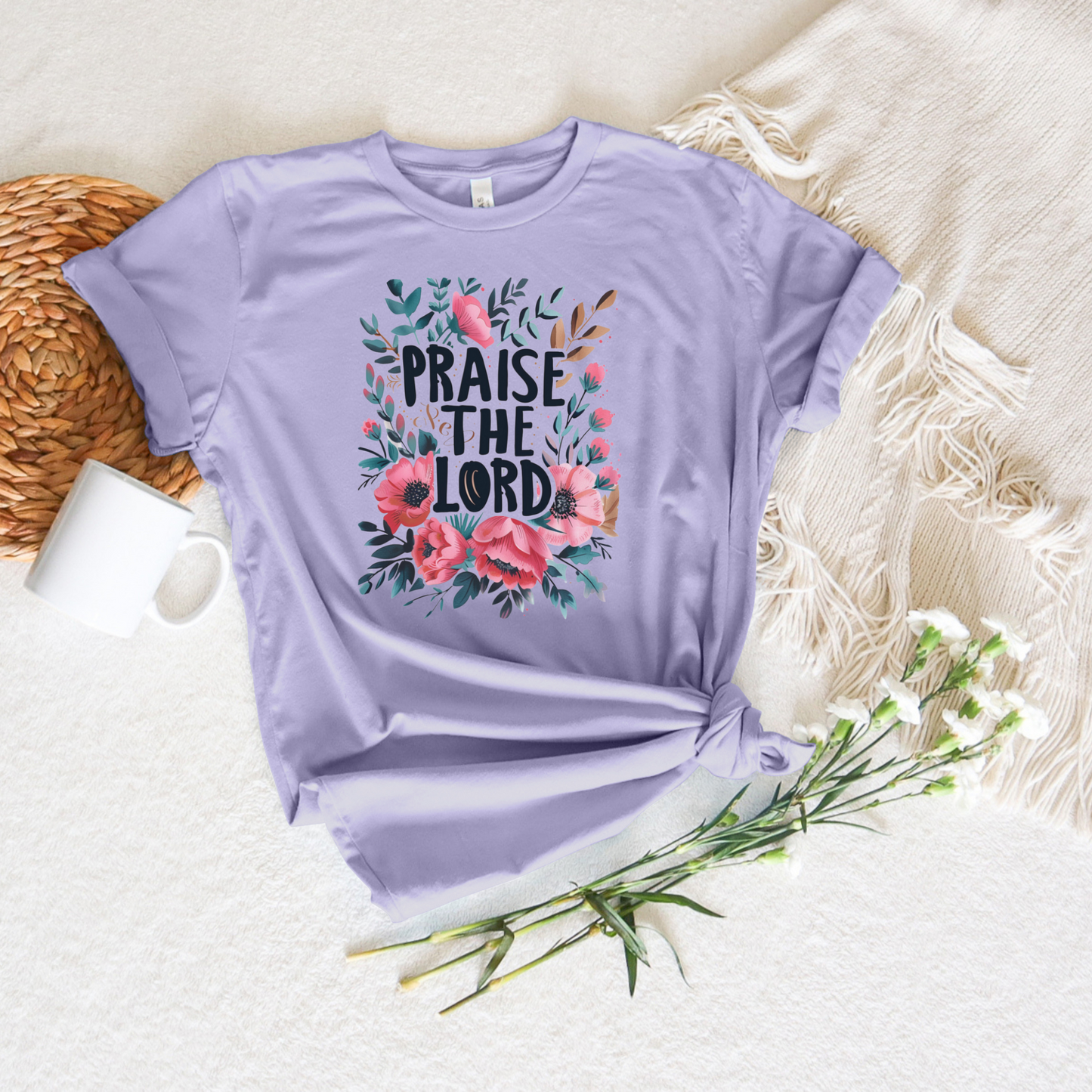 The Praise The Lord Short Sleeve Tee is a lavender T-shirt made from luxurious Airlume cotton. It showcases Praise the Lord text with pink and green floral designs and is displayed on beige fabric next to a woven placemat, white mug, and delicate white flowers.