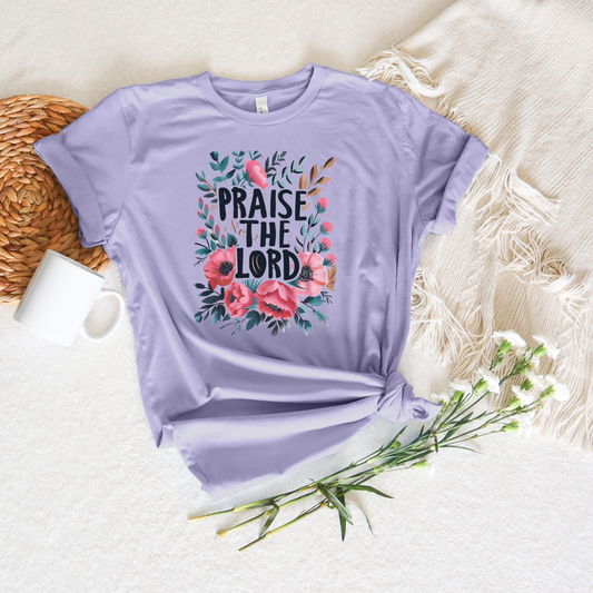 The Praise The Lord Short Sleeve Tee is a lavender T-shirt made from luxurious Airlume cotton. It showcases Praise the Lord text with pink and green floral designs and is displayed on beige fabric next to a woven placemat, white mug, and delicate white flowers.