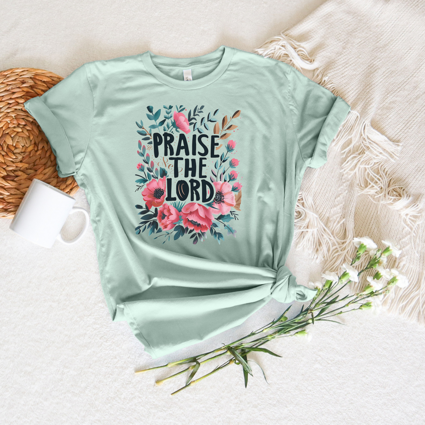 The light green Praise The Lord short sleeve tee, made from buttery soft Airlume cotton, features colorful floral designs. Its placed on a textured white surface beside a fringed fabric, mug, and delicate white flowers.