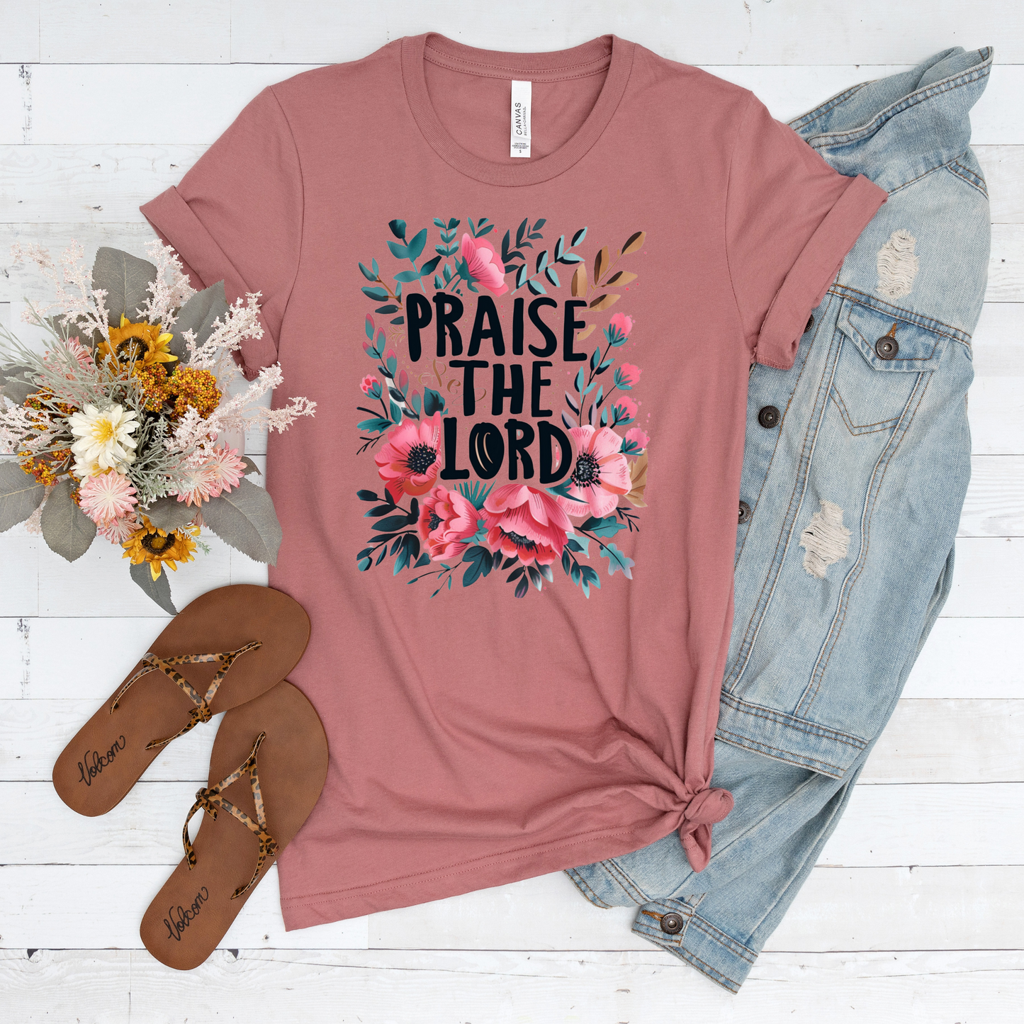 The Praise The Lord Short Sleeve Tee in pink, made from buttery soft Airlume cotton with a floral design, is beautifully arranged with a denim jacket, brown sandals, and a bouquet of flowers on a white wooden surface.