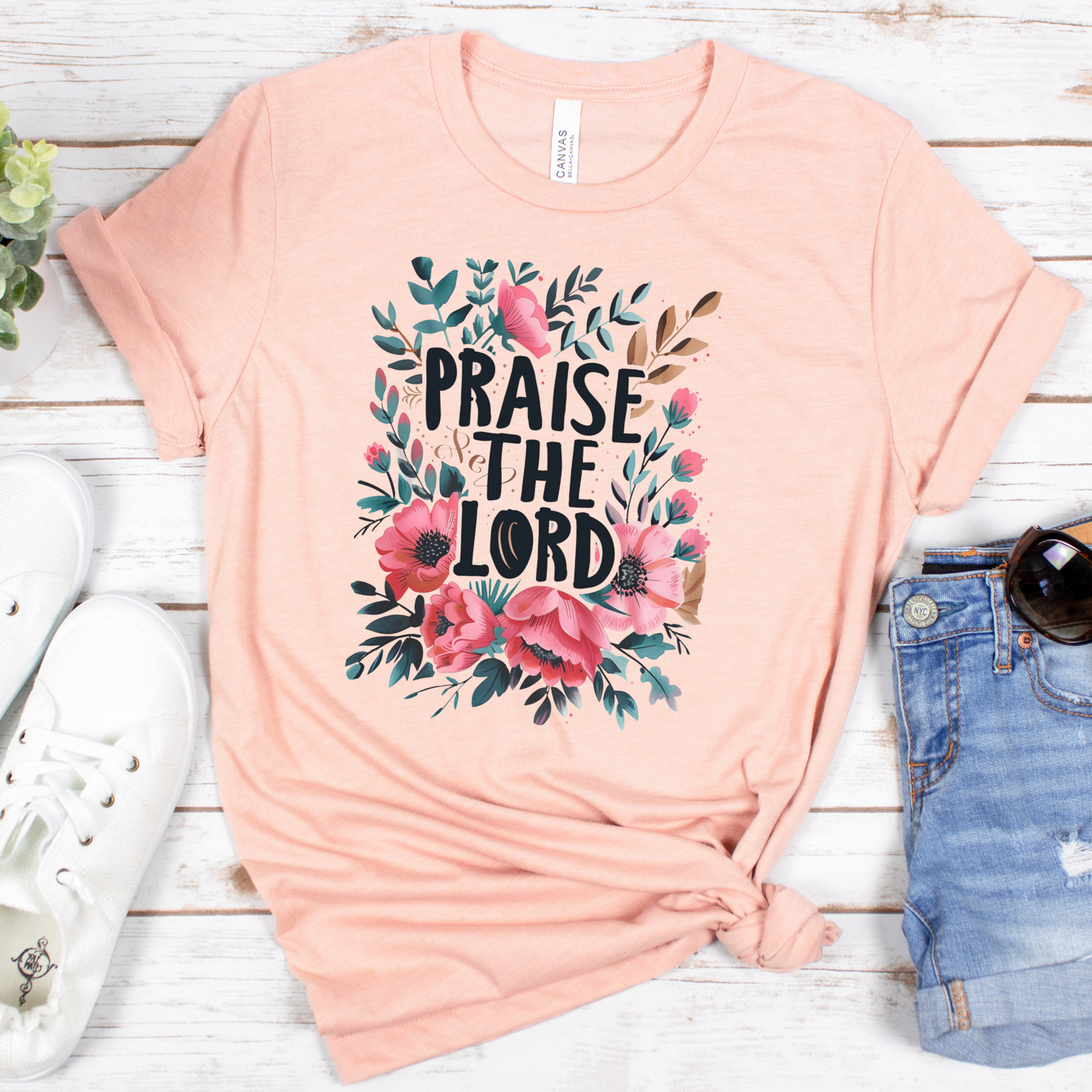 The Praise The Lord Short Sleeve Tee in peach, made from soft Airlume cotton, boasts a floral design of pink and red flowers with green leaves. Styled on a white wooden surface, its ideally matched with white sneakers, denim shorts, and sunglasses for a breezy look.