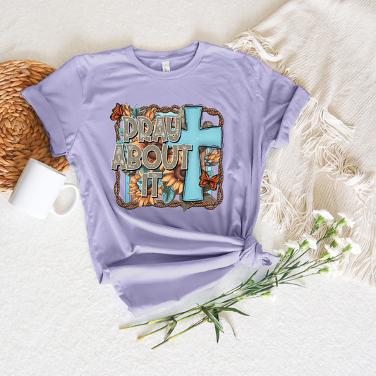 The Pray About It Short Sleeve Tee is a buttery soft lavender T-shirt made from Airlume cotton, featuring colorful PRAY ABOUT IT text, a blue cross, orange flowers, and a brown rope pattern. Its shown on a textured surface with a fringed blanket, white mug, and white flowers.