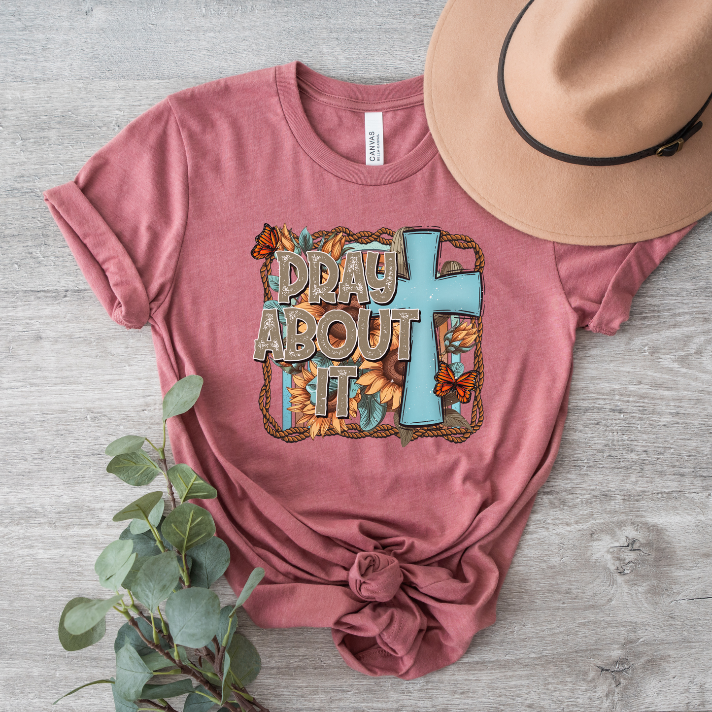 The Pray About It Short Sleeve Tee is made from buttery soft Airlume cotton in pink, featuring Pray About It with a blue cross and floral design. Its styled on light wood with a tan fedora and eucalyptus branch.