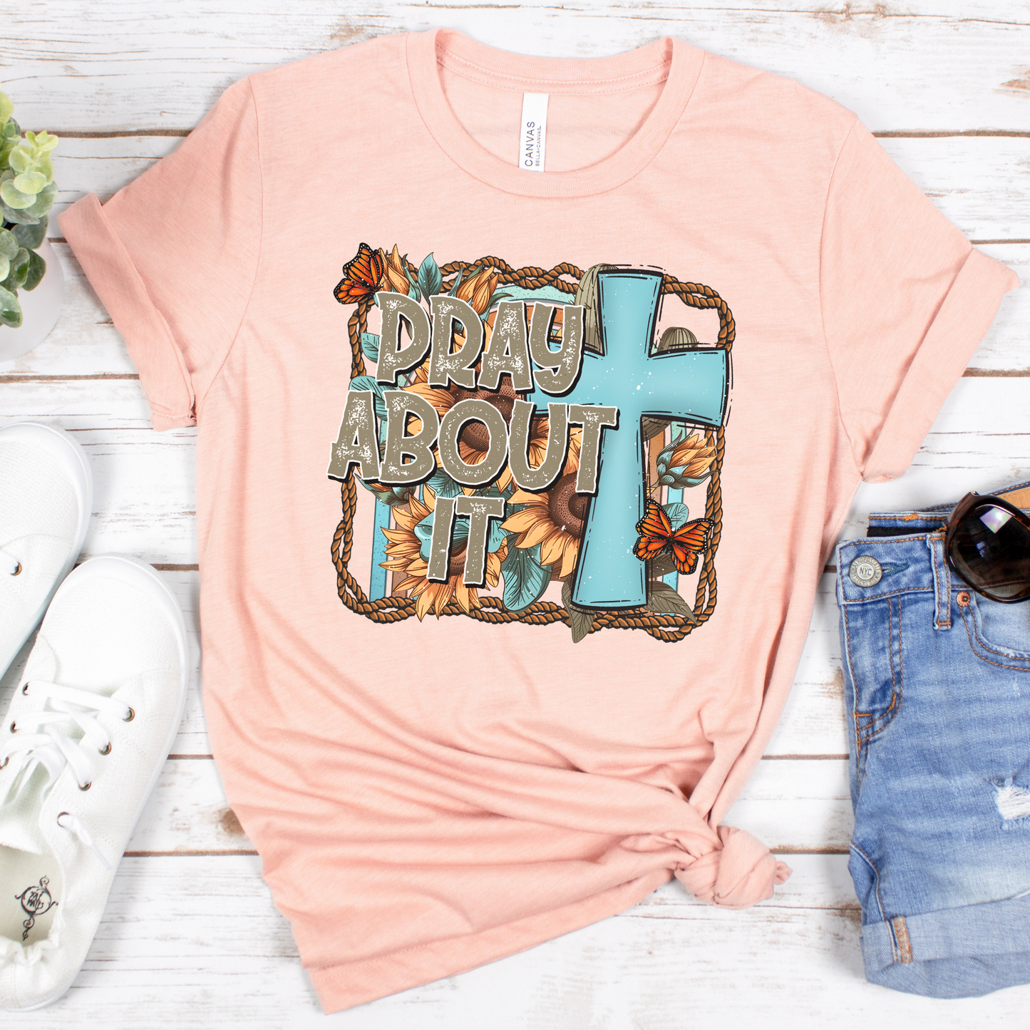 The peach Pray About It Short Sleeve Tee, made from soft Airlume cotton, features a floral-adorned blue cross and text. Styled with white sneakers and denim shorts on a wooden background, its a divine comfort choice for any day.