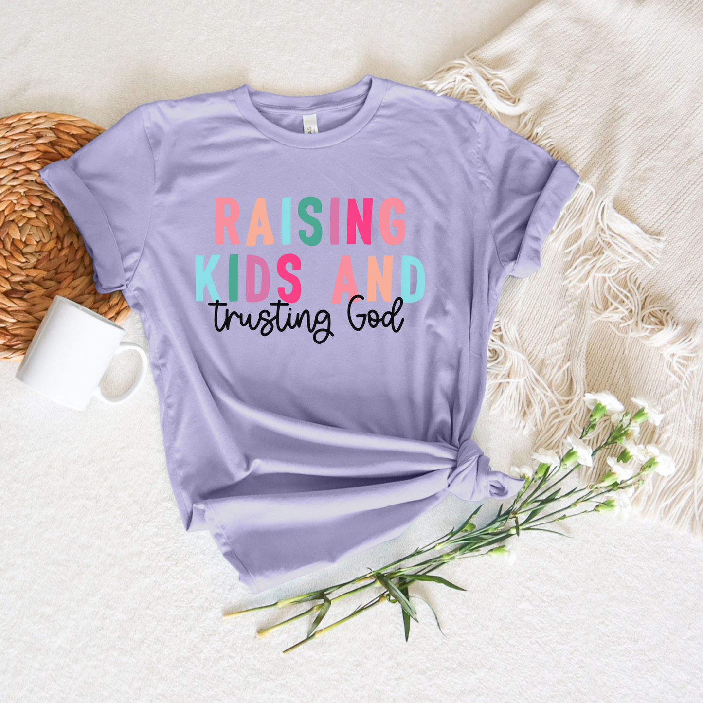 The Raising Kids and Trusting God Short Sleeve Tee in lavender, crafted from soft Airlume cotton, features the colorful phrase against a cream woven blanket, accompanied by a white mug, wicker mat, and delicate white flowers.
