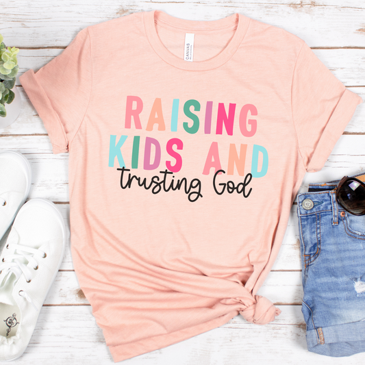 The Raising Kids and Trusting God Short Sleeve Tee, in peach and made from buttery soft Airlume cotton, displays vibrant text. Its neatly folded beside white shoes and blue denim shorts on a light wooden surface, accompanied by a small plant and sunglasses.