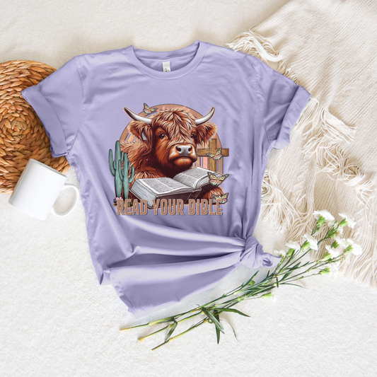 The Read Your Bible Highland Cow Short Sleeve Tee, made from soft Airlume cotton, showcases a Scottish Highland cow, open Bible, and cactus illustration with the text READ YOUR BIBLE. Presented on a beige blanket with flowers and paired with a mug and coaster. Ideal for those who value faith and comfort.