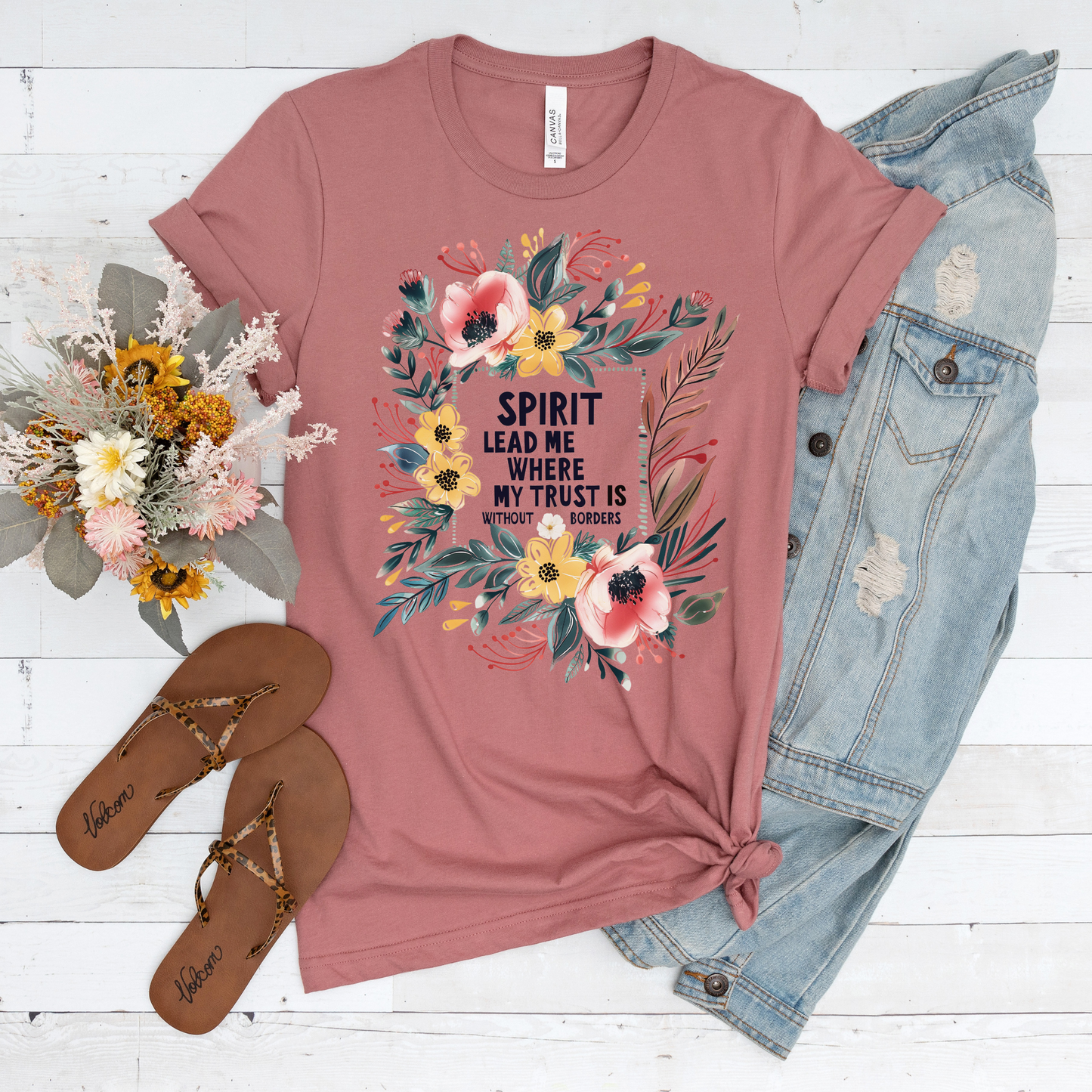 The Spirit Lead Me Pink Floral Short Sleeve Tee is made from buttery soft Airlume cotton, featuring a floral design and the phrase Spirit lead me where my trust is without borders. Its styled with a denim jacket, brown sandals, and flowers on a white wooden surface. Crafted with God in mind.