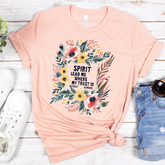 The Spirit Lead Me Pink Floral Short Sleeve Tee, made from Airlume cotton, is buttery soft and features a pink floral design encircling the text: Spirit lead me where my trust is without borders. Its styled on a white wooden surface with white sneakers, a small plant, and denim shorts.