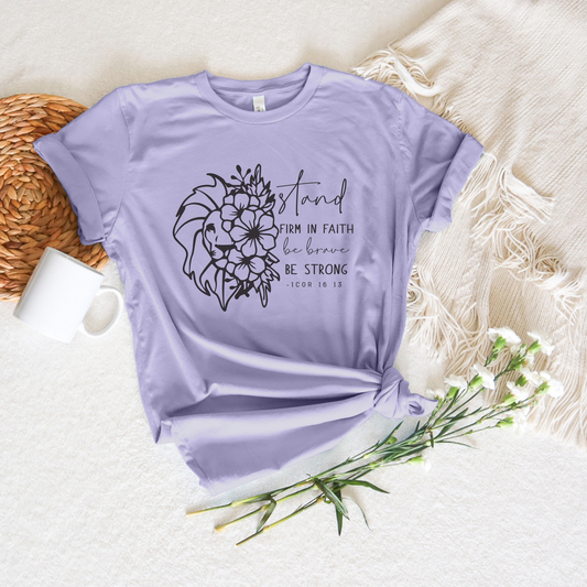 The Stand Firm In The Faith Short Sleeve Tee in lavender showcases a floral lion and Stand firm in faith, be brave, be strong - 1 COR 16:13. Its made from buttery soft Airlume combed cotton, styled alongside a beige blanket, white mug, wicker placemat, and delicate white flowers.