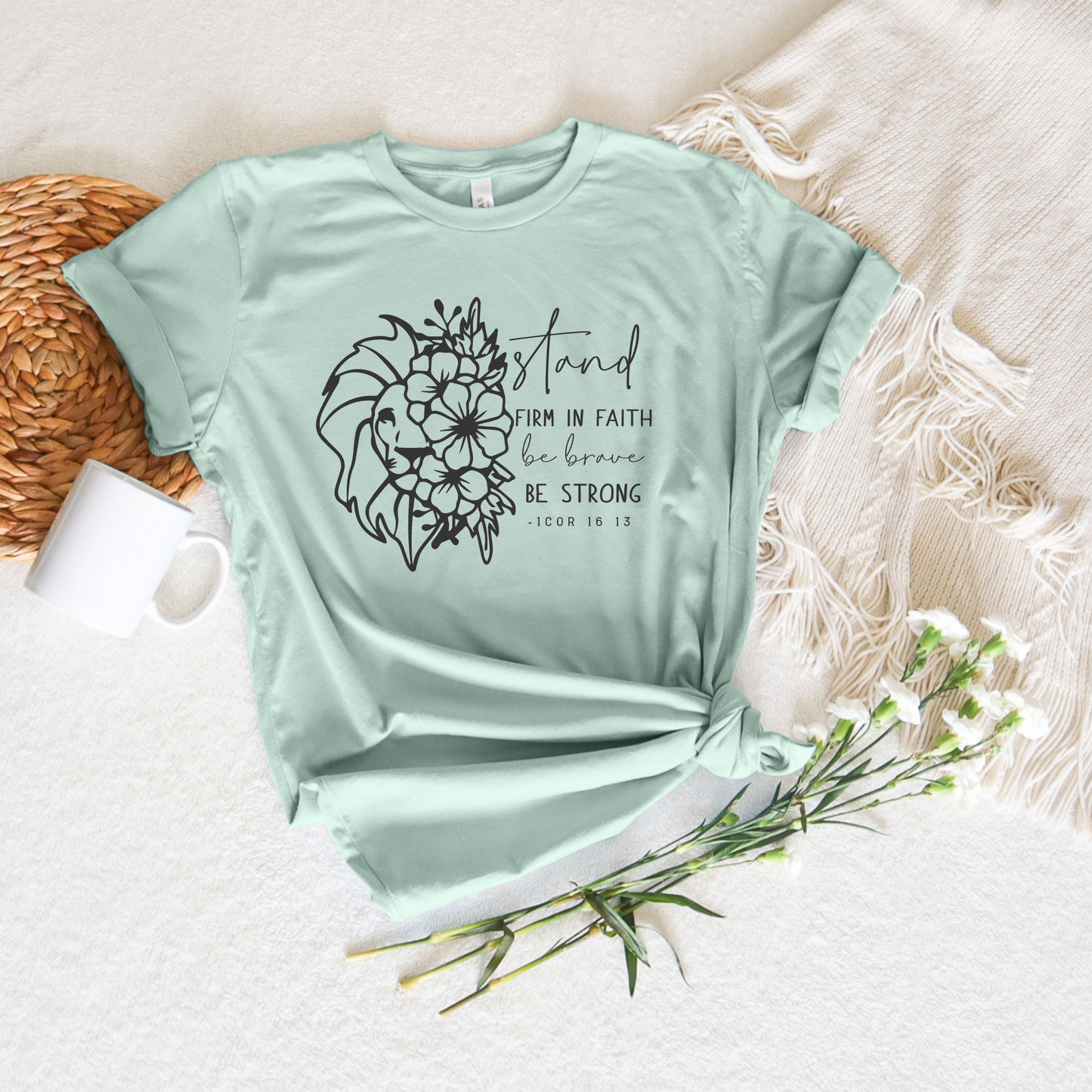 The Stand Firm In The Faith Short Sleeve Tee, in light green and made of buttery soft Airlume combed cotton, features floral and lion head artwork with the text Stand firm in faith, be brave, be strong - 1 Cor 16:13, displayed on a white blanket with a wicker placemat, white mug, and flowers.