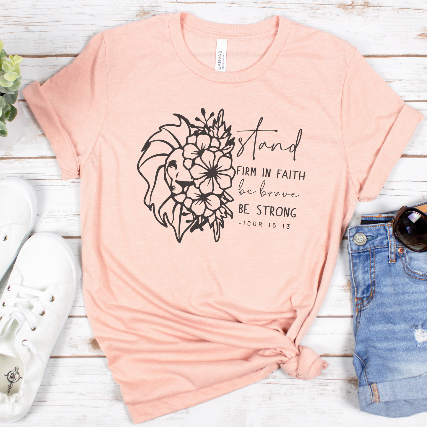 The Stand Firm In The Faith Short Sleeve Tee, in peach, crafted from buttery soft Airlume combed cotton with a floral lion and Stand firm in faith, be brave, be strong. -1 Cor 16:13 text, is styled with rolled-up blue denim shorts and white sneakers on a white wooden surface with a potted plant.