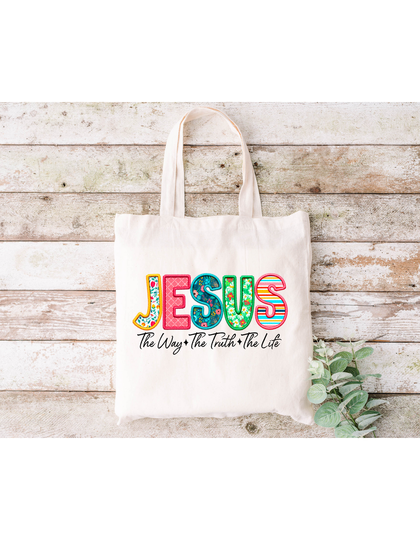 The Jesus The Way The Truth The Life Canvas Bag is a beige cotton tote with Jesus in vibrant letters, above the phrase The Way, The Truth, The Life. Its perfect for Bible study and is presented on a rustic wooden background with green leaves in the bottom right corner.